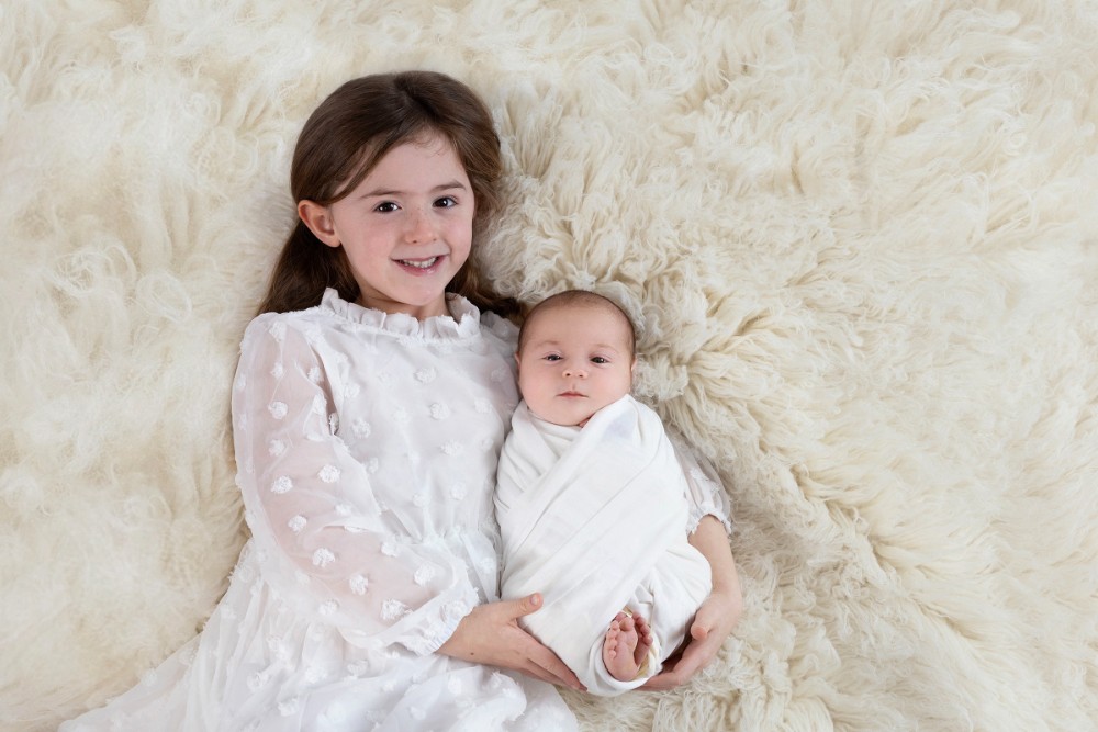 Big sister cuddling newborn sibling on a fluffy white rug, a heartwarming family moment captured in Maldon, Essex